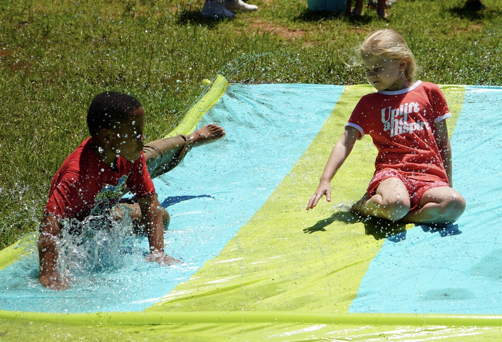 Students slide down a slip n slide on grass outside a school.