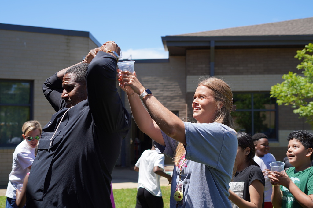 A principal and a teacher play a game outdoors. The principal pours water over his head and into a cup the teacher is holding behind him. 