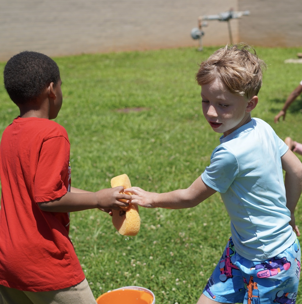 A boy hands another boy a sponge while they play a water relay race outside on grass.