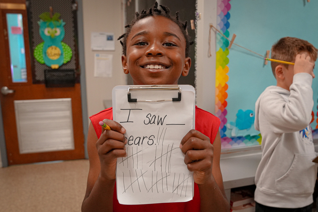 A student smiles and stands in a school hallway. He holds up a piece of paper marking off how many bears he saw on a scavenger hunt.