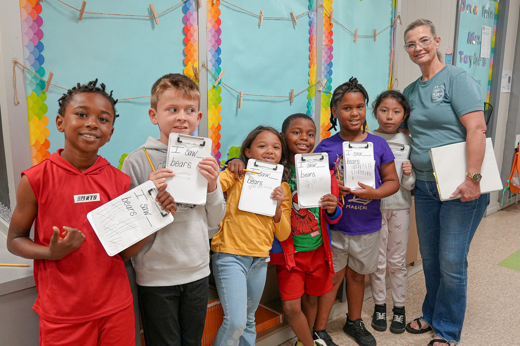 Students hold up pieces of paper marking off how many bears they saw on a scavenger hunt. They stand with their teacher in a school hallway.
