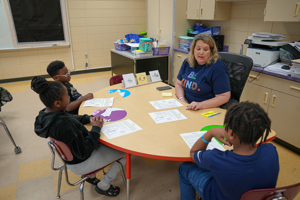 A teacher sits at a table with students inside a classroom and leads a lesson.