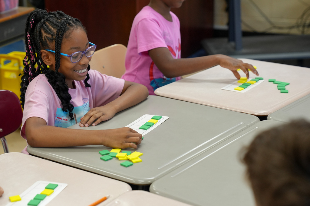 A student smiles while doing an assignment in a classroom. She places green and yellow tiles onto a piece of paper.