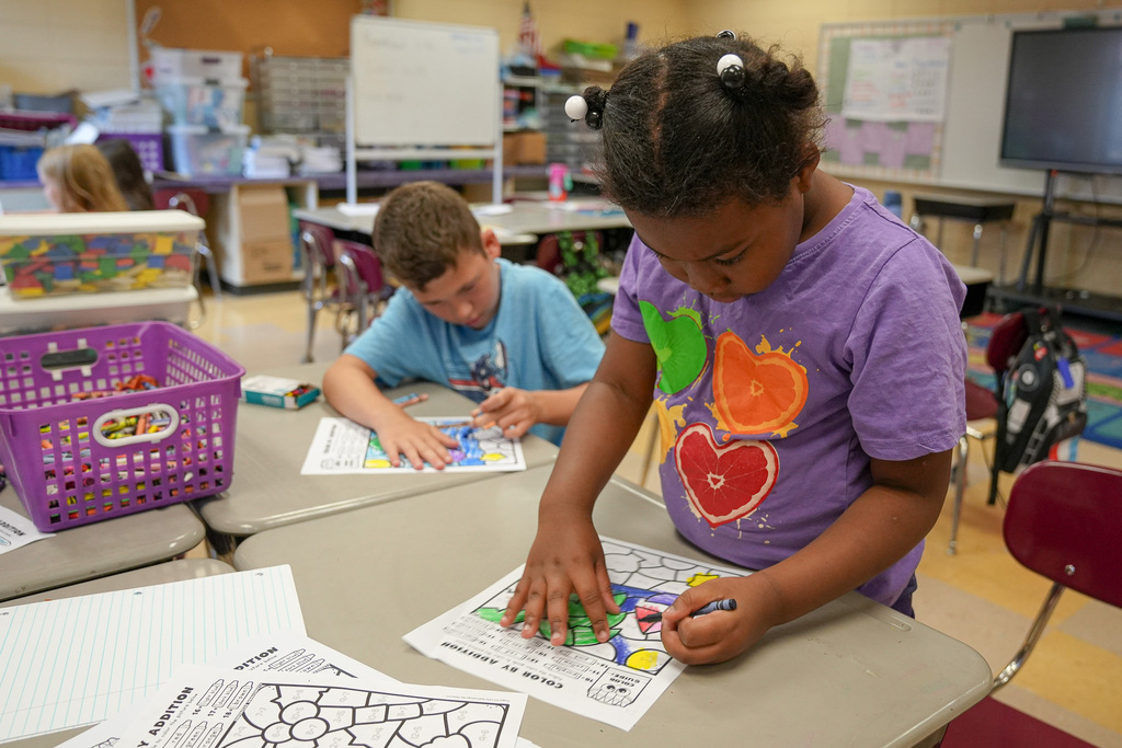 A student colors on a sheet of paper while standing at a desk inside a classroom.