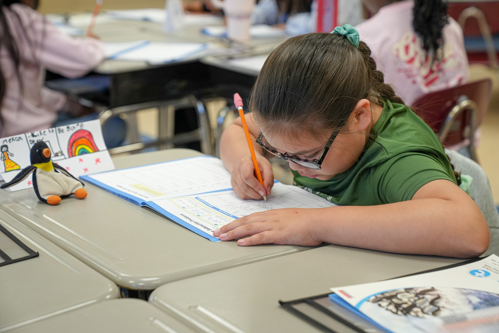 A student writes in a workbook while sitting at a desk inside a classroom.