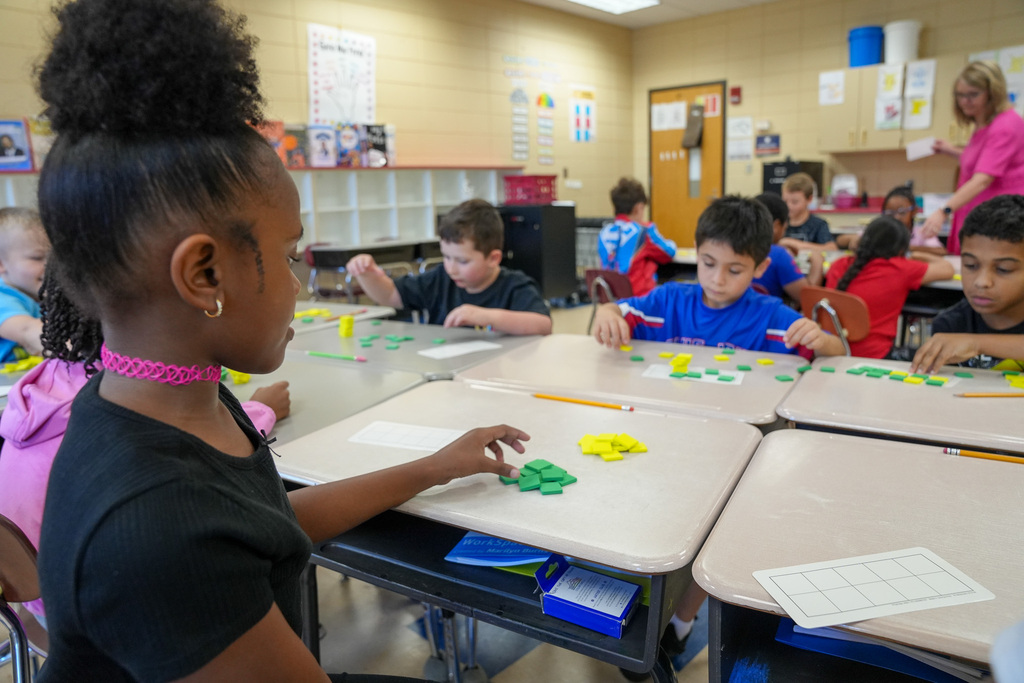 Students sit at desks inside a classroom and place colorful tiles on a piece of paper.