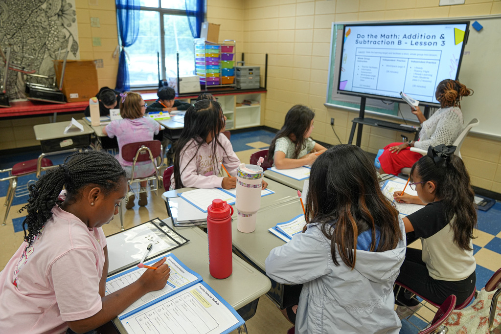 A teacher leads a lesson at the front of a classroom. Students listen while sitting at desks.