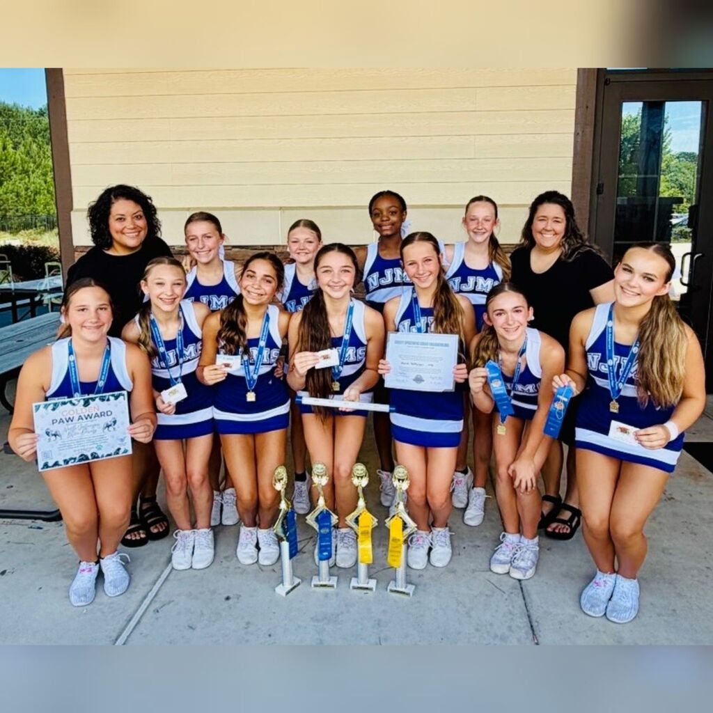 The picture is of the NJMS Cheer Team in their blue and white uniforms. They are posing in two rows with 4 trophies at the bottom of the first row and some girls are holding certificates or medals. There are 7 girls in the front row and 4 girls in the back row with a coach on either side of the four girls.