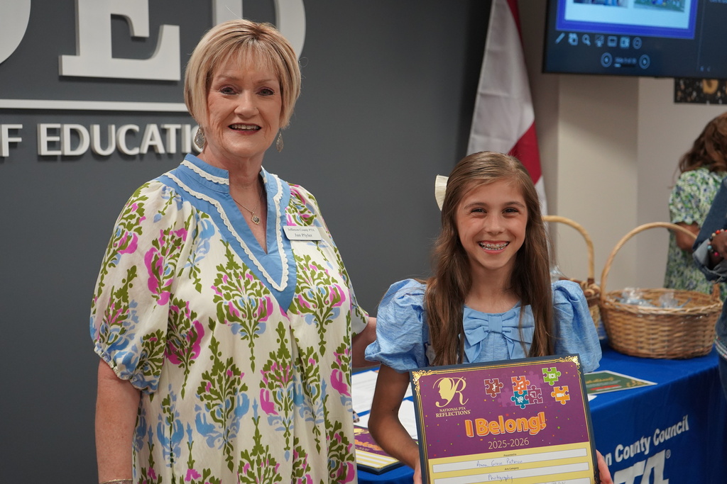 A woman and a girl smile and pose for a photo inside a room in the Jefferson County Schools Board of Education. The girl holds a certificate in her hands. 