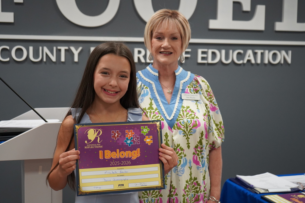 A woman and a girl smile and pose for a photo inside the Jefferson County Board of Education. The girl holds up a certificate.