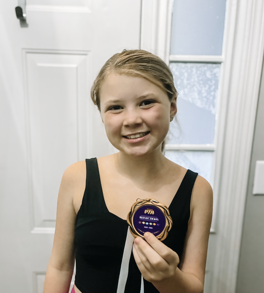 A girl smiles and holds up a medal that says "PTA Reflections" while standing in front of a door inside a building.