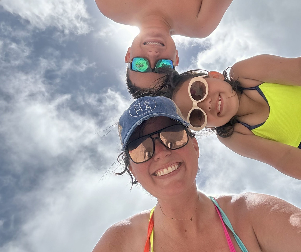 A woman, a girl, and a boy take a selfie in front of a blue sky. They wear swimsuits and smile.