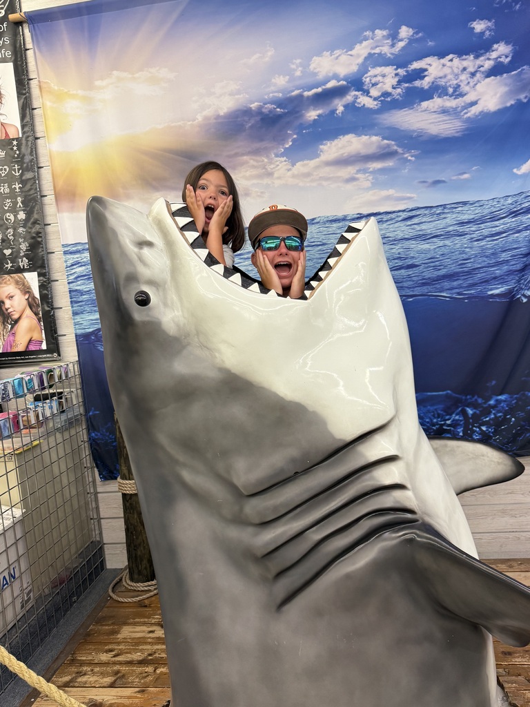 Two children pose for a photo behind a shark prop in a building. They make silly faces pretending to be scared.