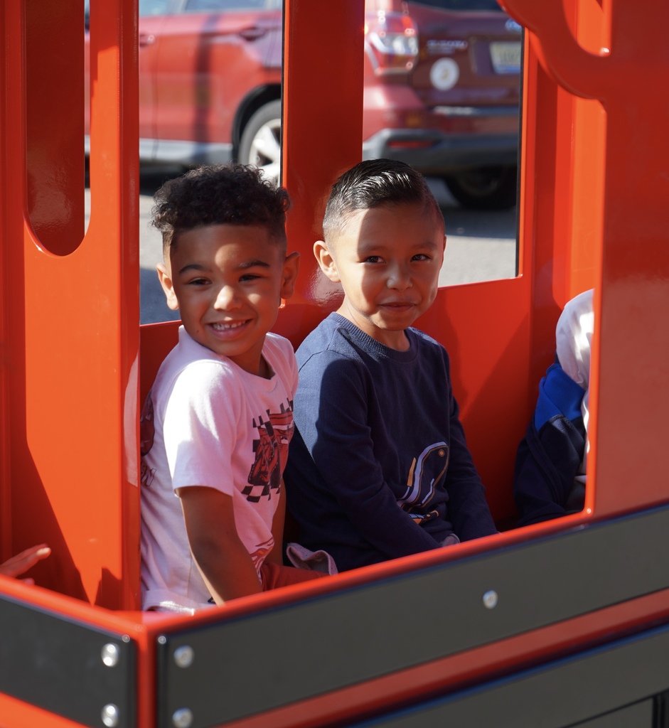 Students smile while riding inside a red carnival-style train inside a school parking lot.