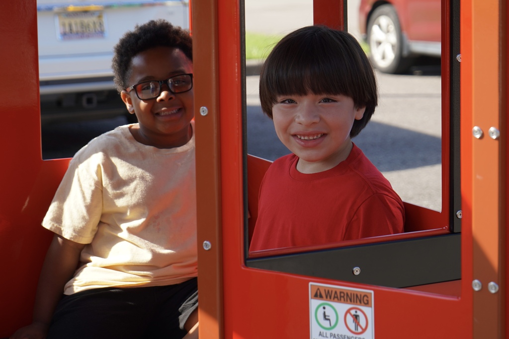Two students smile while sitting in a red train car inside a school parking lot.