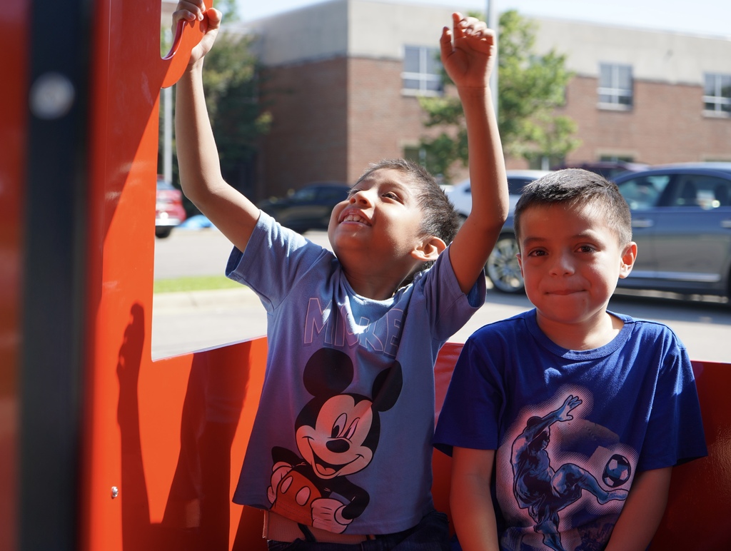 Students smile while riding a carnival-style train. One of the students raises his arms in the air.