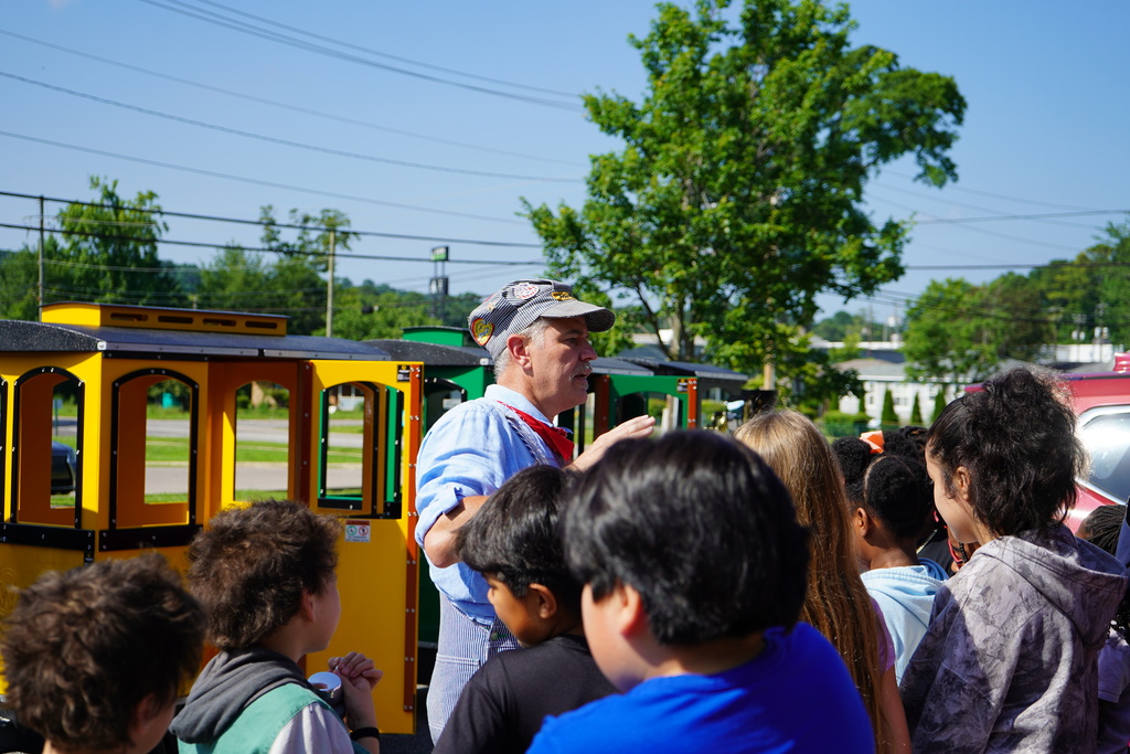 A man speaks to a group of students while standing outside a colorful train. He wears a conductor costume.