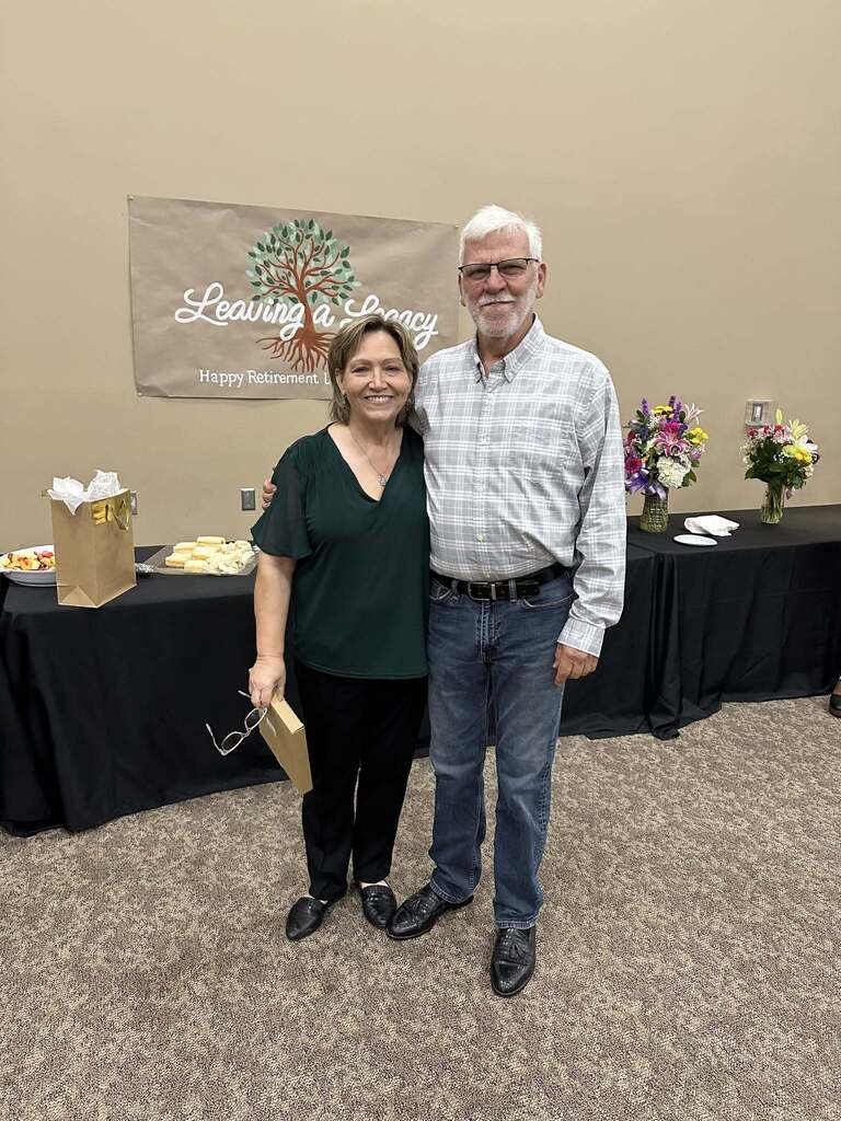 A woman and a man stand together for a photo. They stand in front of tables inside a room.