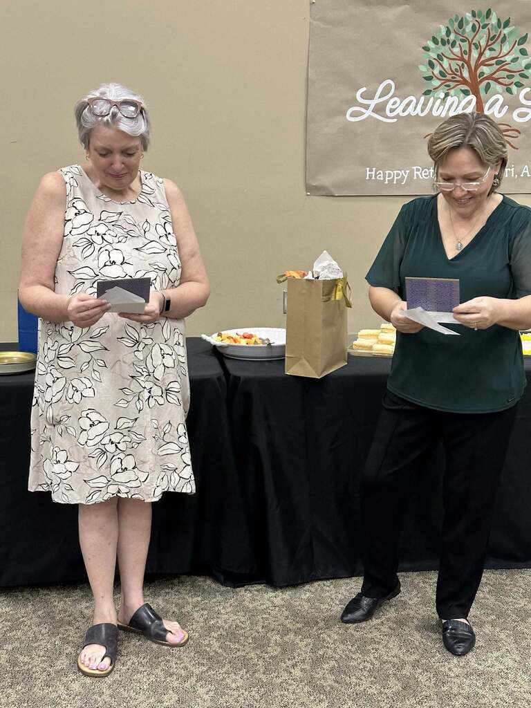 Two women stand in a room in front of tables and a hand-painted sign. They read cards during an event celebrating their retirement.