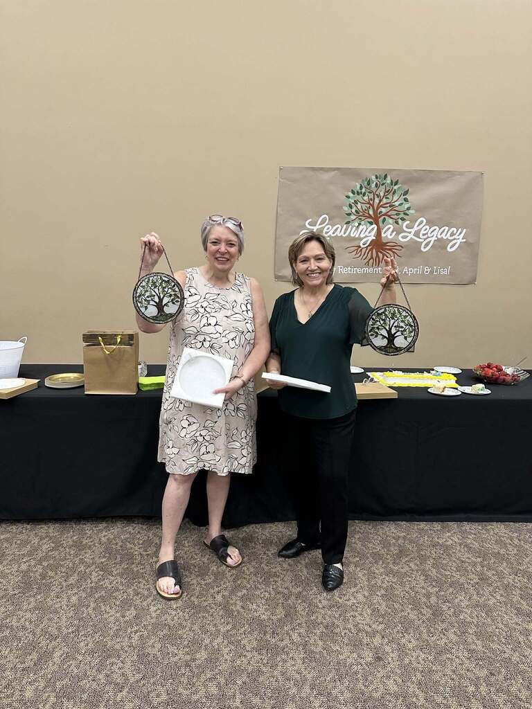 Two women stand together in a room in front of tables and a hand-painted sign. They each hold up tree-themed art work.