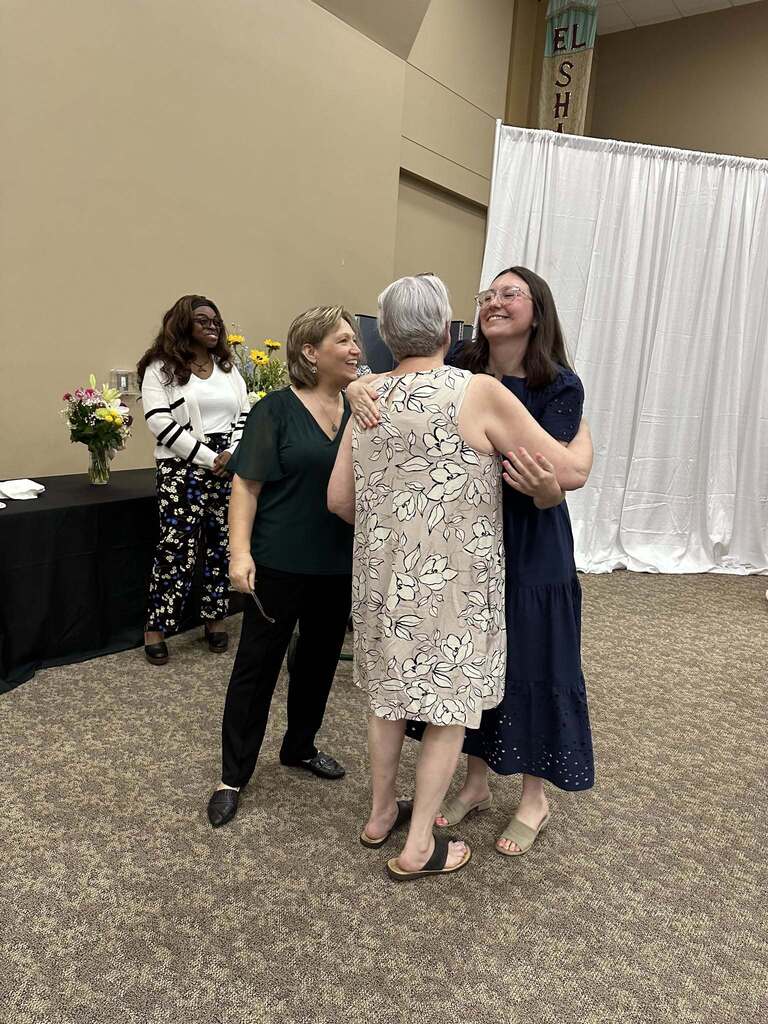 Two women hug each other while standing inside a room. Another women stands close to them and smiles. Behind them, a fourth woman stands next to tables with flowers on them.