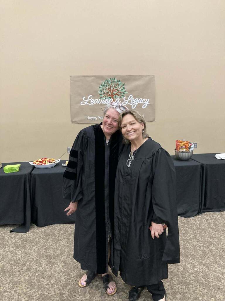 Two women stand together for a photo and smile. The women wear graduation gowns and stand in a room in front of a table with food on it.