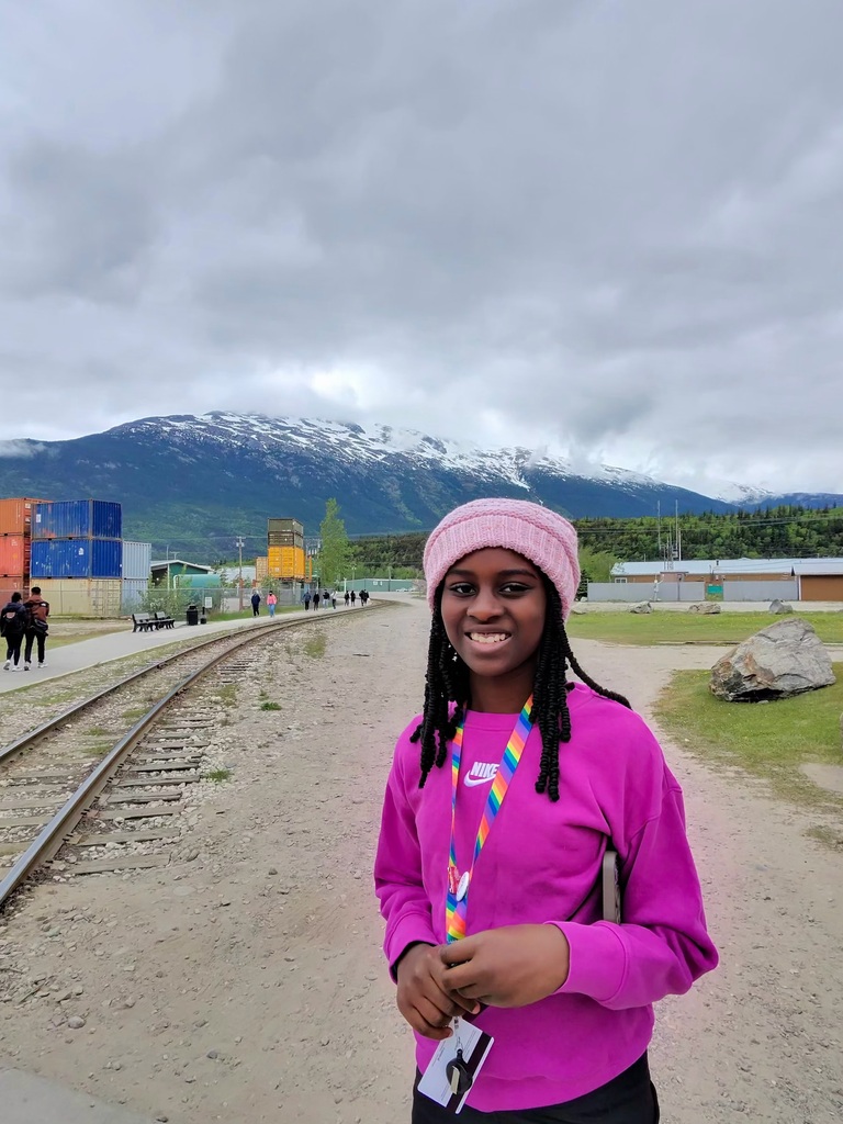 A girl wearing a pink sweatshirt and a pink hat smiles while standing in front of a snow-capped mountain and train tracks.