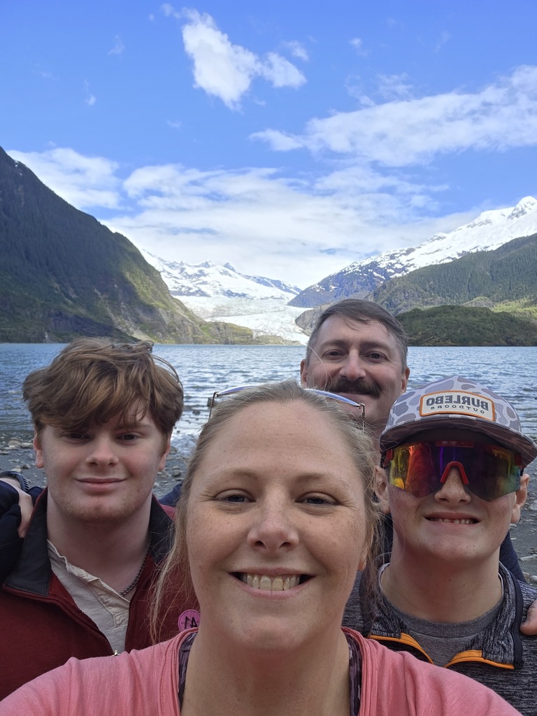 A woman, man, and two children smile for a selfie while standing in front of a large body of water and snow-capped mountains.
