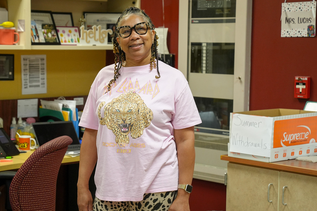 A woman smiles for a photo inside a school office. She wears a Def Lepard shirt and leopard print pants.