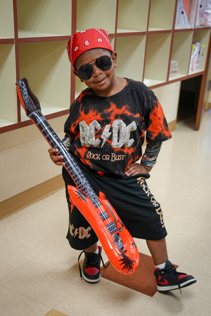 A little boy dressed in a rockstar themed outfit poses for a photo. He smiles and stands in a school while holding a guitar prop.