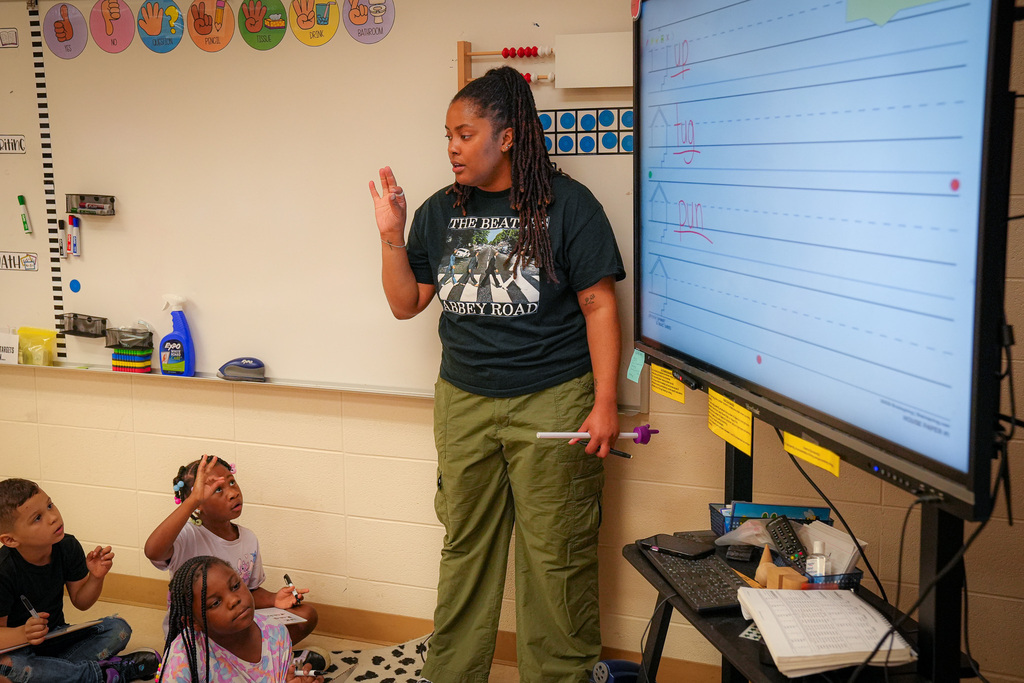 A teacher leads a lesson inside a classroom. Students sit on the floor and listen. The teacher wears a Beatles themed shirt.
