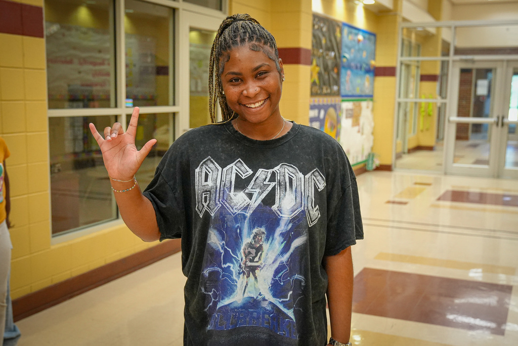 A woman signs "I love you" with her hand and poses for a photo inside a school hallway. She smiles and wears an AC/DC shirt.