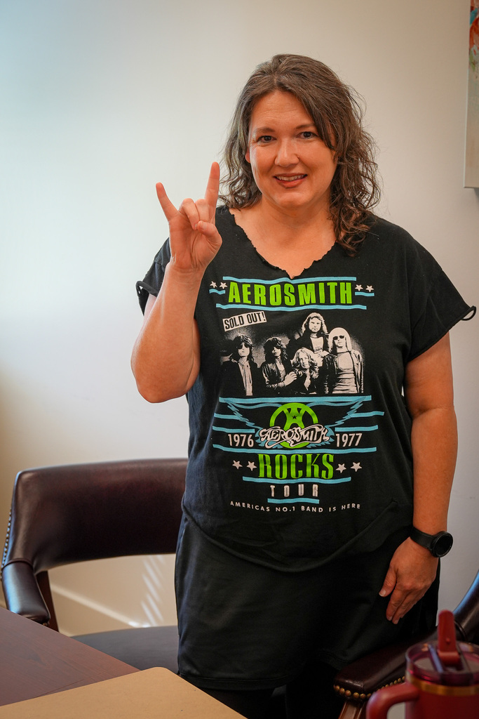 A woman poses for a photo inside a room. She holds up the rock and roll symbol with her hand and wears an Aerosmith shirt.
