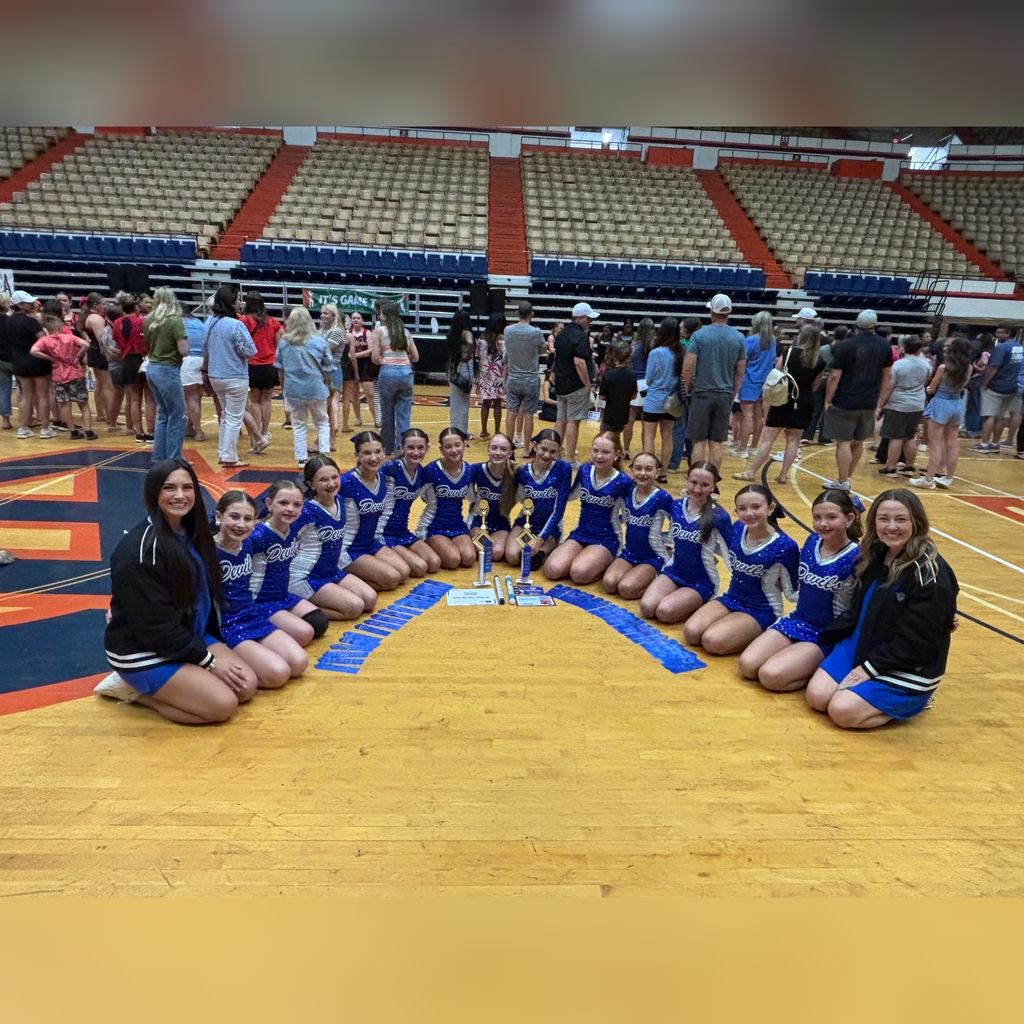 The dance team girls are kneeling in a semicircle with the two coaches on either end. In front of the girls are the ribbons, certificates, and trophies that they won.