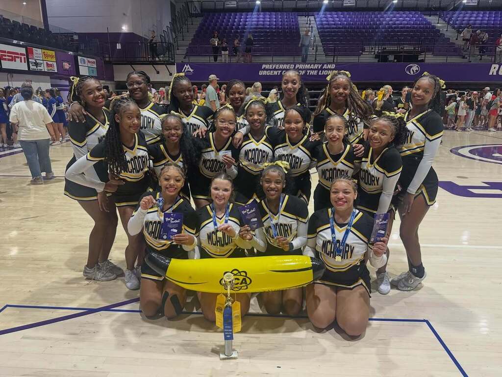 McAdory High School cheerleaders pose for a photo inside a gym. They smile and stand and kneel behind trophies. The cheerleaders in the front of the photo hold a UCA inflatable banana.