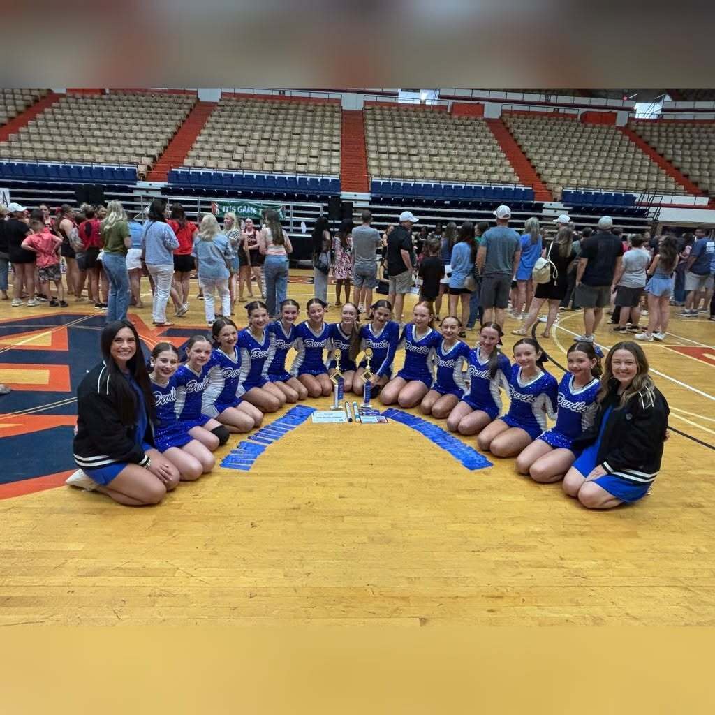 North Jefferson Middle School dance team members pose for a photo inside a gym. They kneel around trophies and awards displayed on the floor.