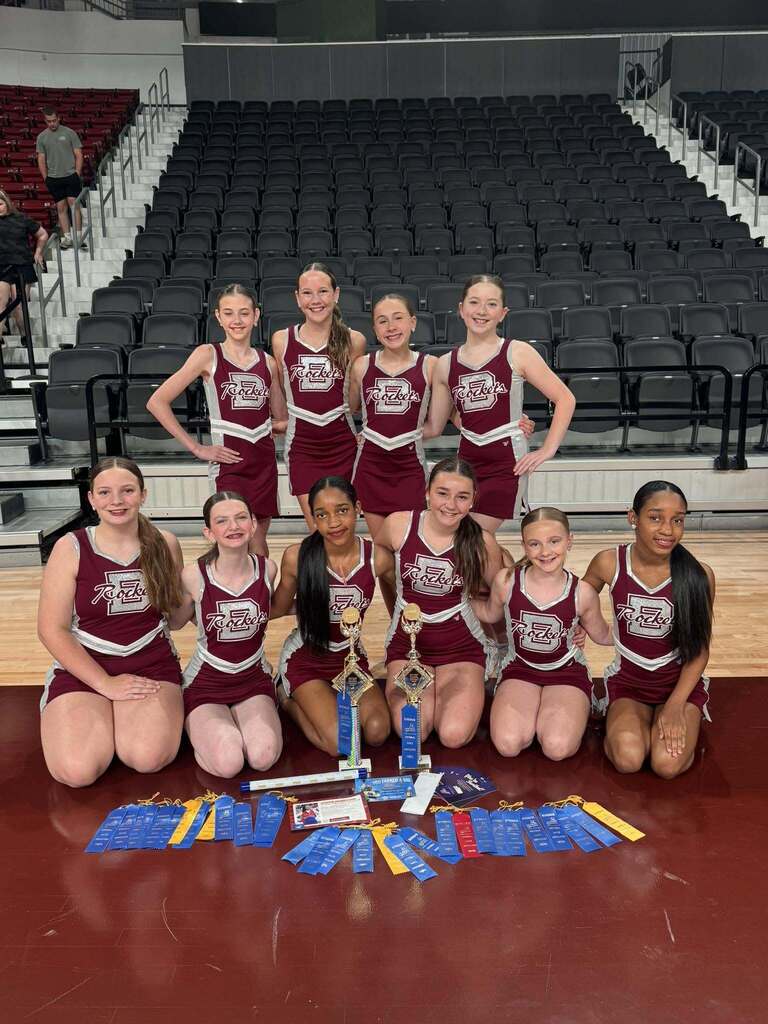 Bragg Middle School dance team members smile for a photo inside a gym. They stand and kneel behind awards that are sitting on the ground.