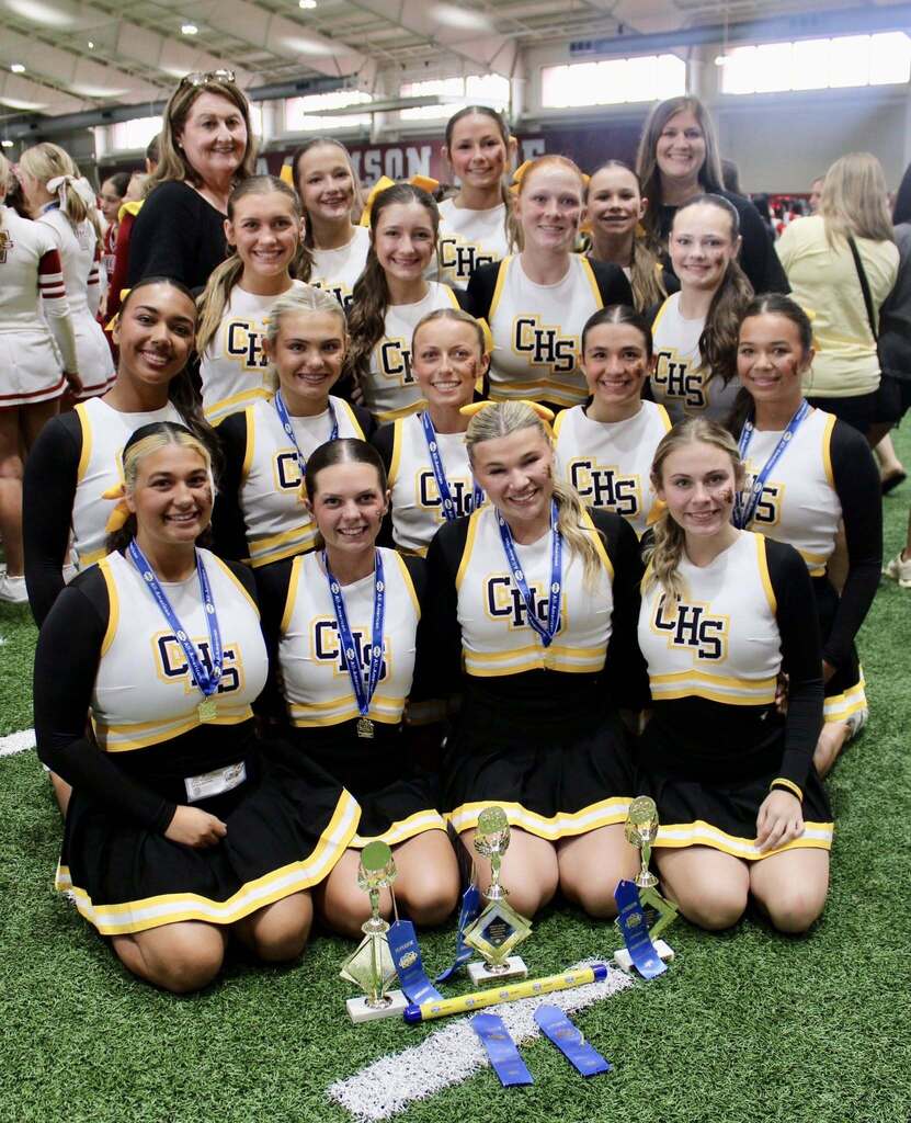 Corner High School cheerleaders pose for a photo inside a gym. They stand and kneel on grass. Awards sit on the ground in front of the cheerleaders.