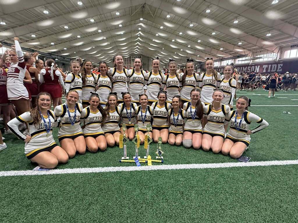 Corner Middle School cheerleaders pose for a photo inside a gym. They stand and kneel on turf and pose behind trophies.