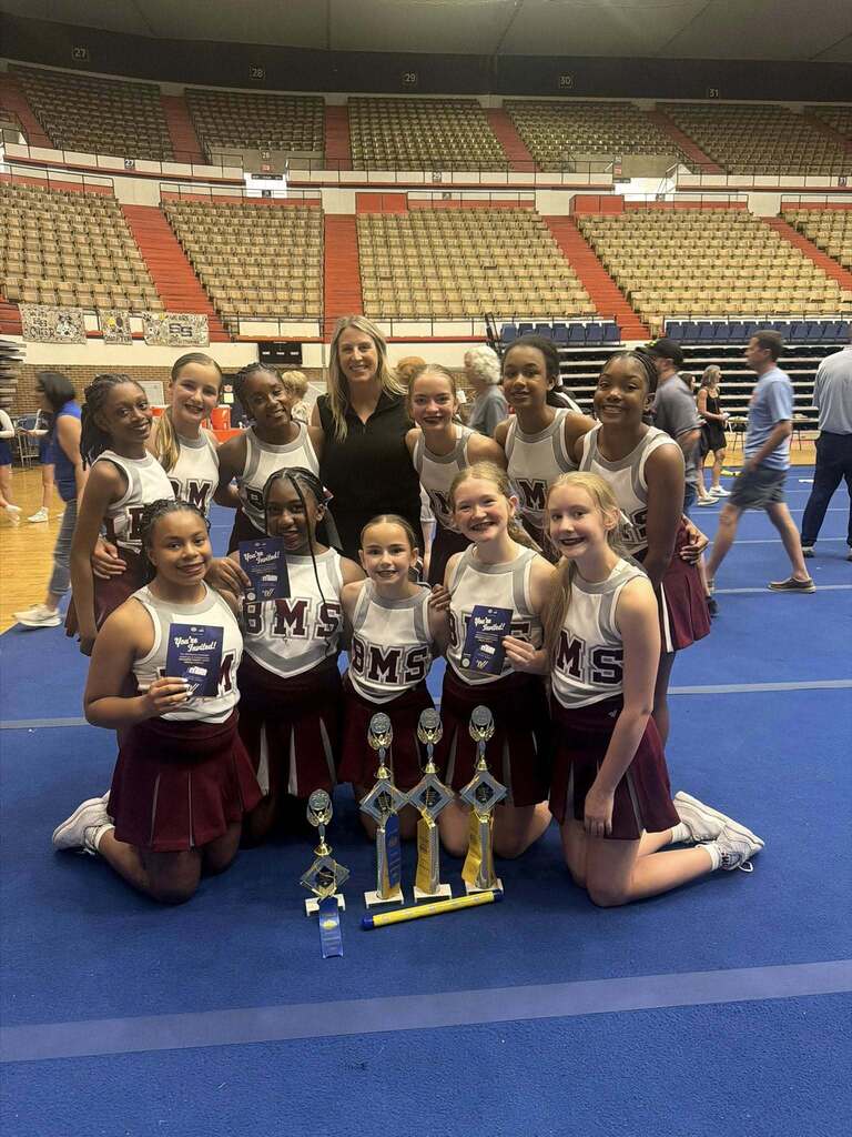 Bragg Middle School cheerleaders pose together for a photo on a blue cheer mat. They stand and kneel next to trophies.