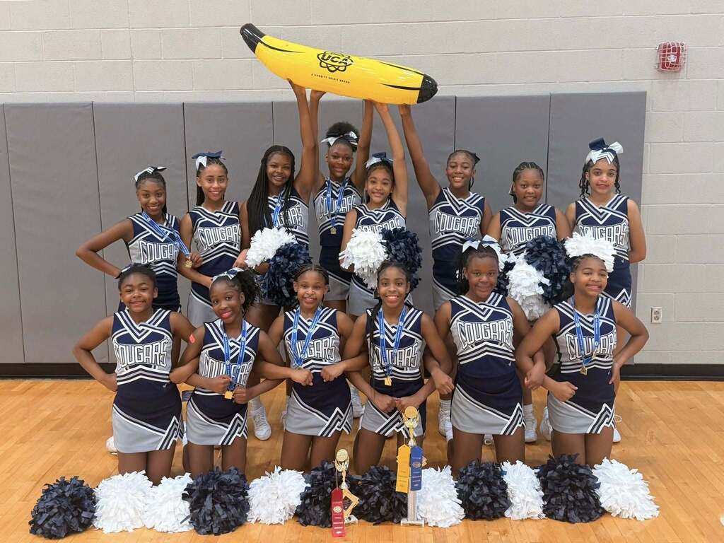 Clay-Chalkville Middle School cheerleaders pose for a photo inside a gym. They stand and kneel behind awards that are sitting on the ground. The dancers in the back of the photo hold up an inflatable UCA banana.