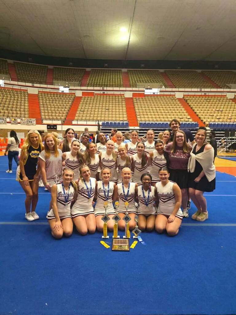 Gardendale cheerleaders pose for a photo inside a gym. They smile and pose behind awards that are sitting on the ground. The cheerleaders in the front of the photo wear medals around their necks.