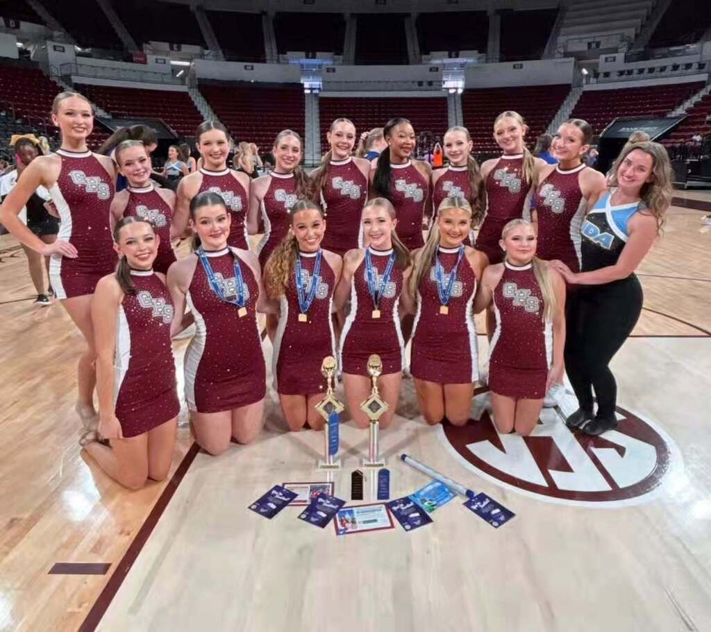 Gardendale High School dance team members pose for a photo inside a gym. They wear sparkly maroon uniforms and pose behind trophies and other awards. Some of the dancers wear medals around their necks.