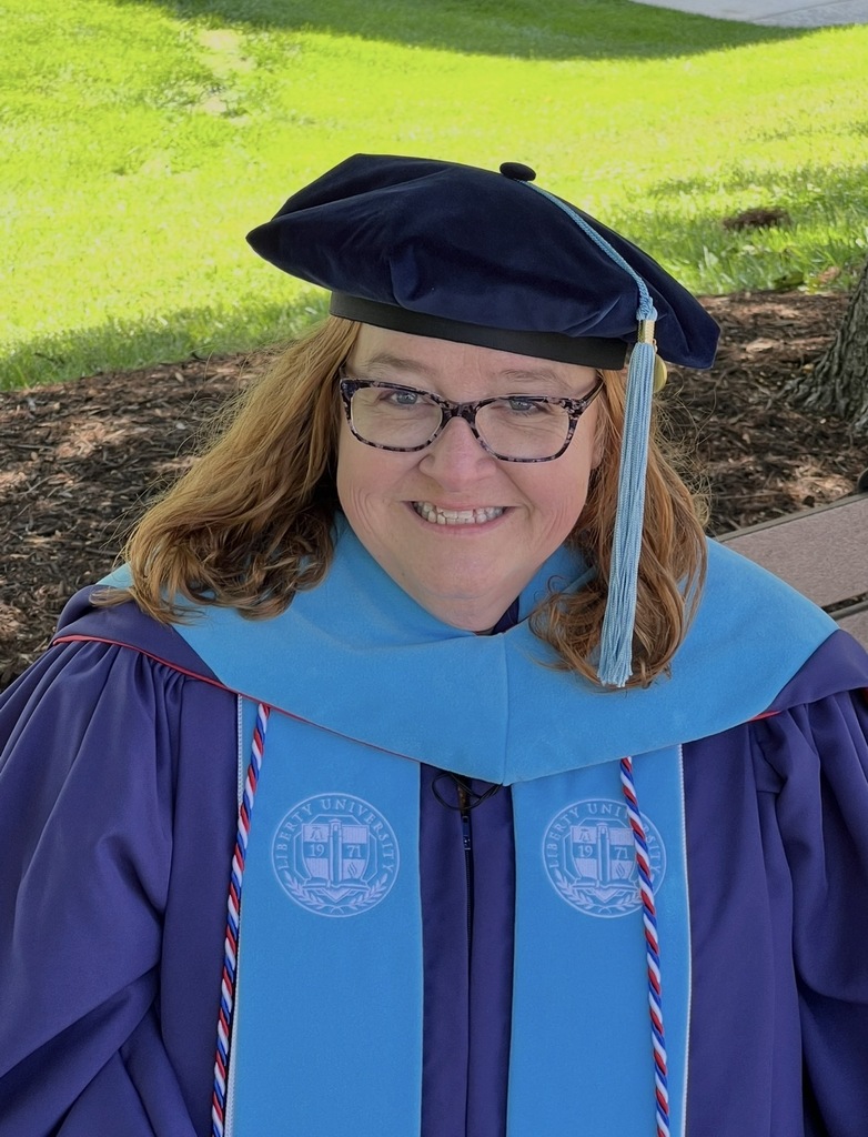 Jennifer Akins Moore smiles for a photo while sitting outdoors. She wears a graduation gown and cap.