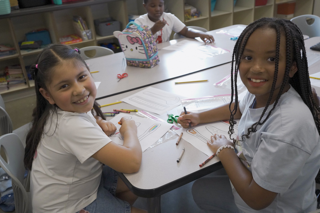 Two girls sit at a table and smile. They color on pieces of paper.