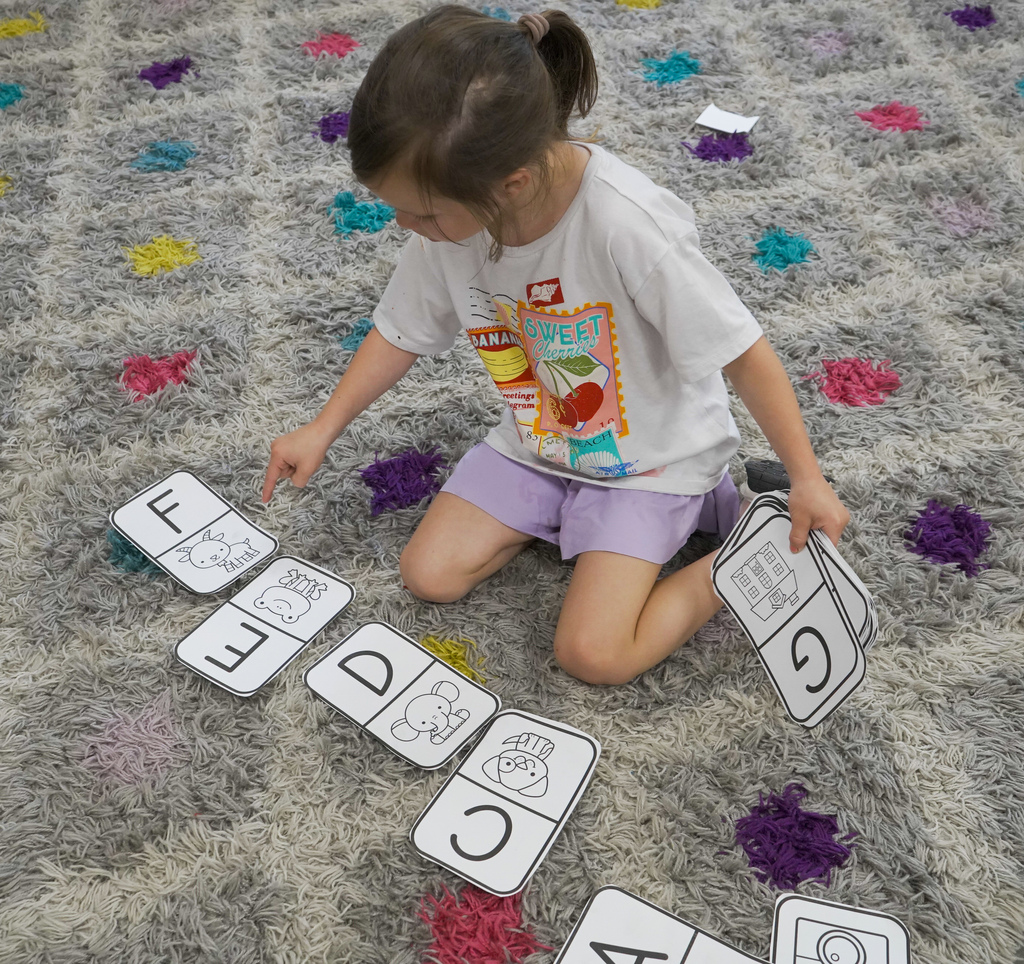 A child sits on a carpet and picks up pieces of paper that are sitting on the ground. The papers have letters on them.