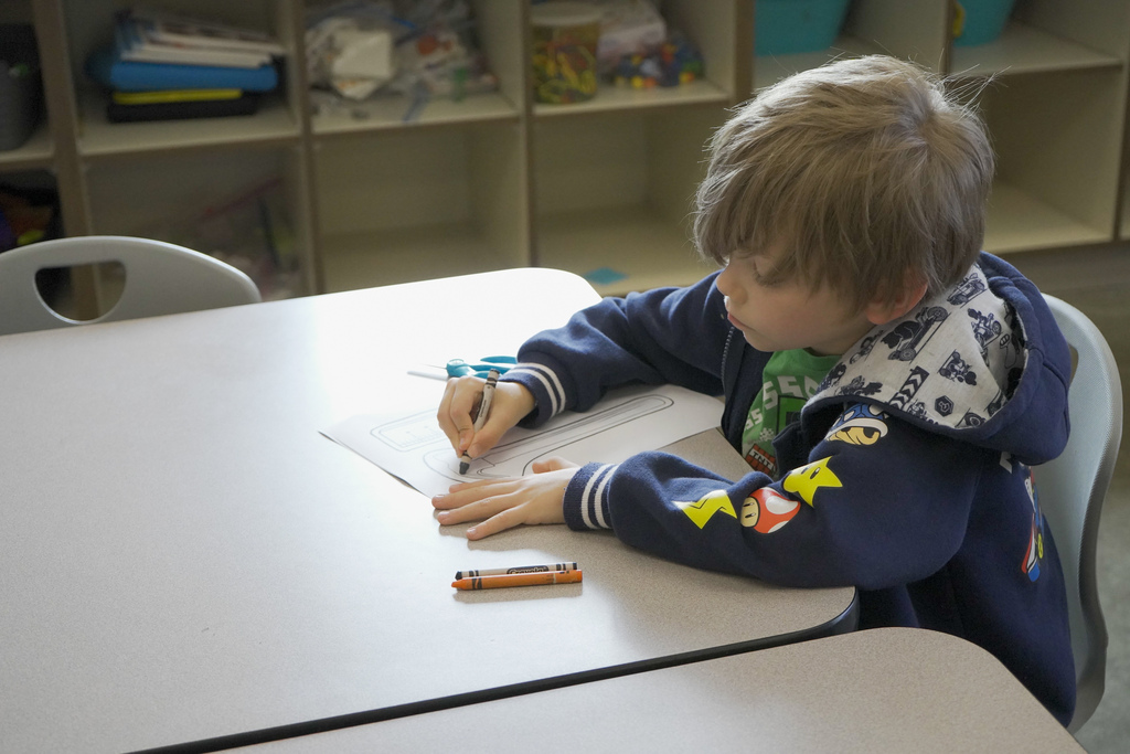A child sits at a table inside a classroom and colors on a piece of paper.