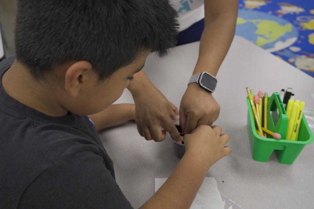 A teacher helps a student plant a seed inside a classroom.
