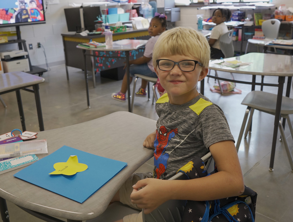A student sits at a desk in a classroom and smiles. The student wears a Spiderman shirt and has pieces of paper and supplies on his desk.