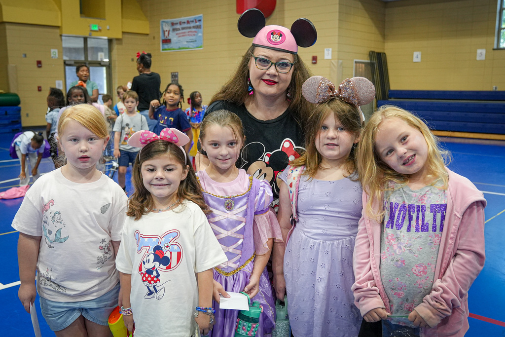 A group of girls wearing Disney-themed outfits smile for a photo inside a school gym. They pose with an educator who wears a Disney-themed hat.